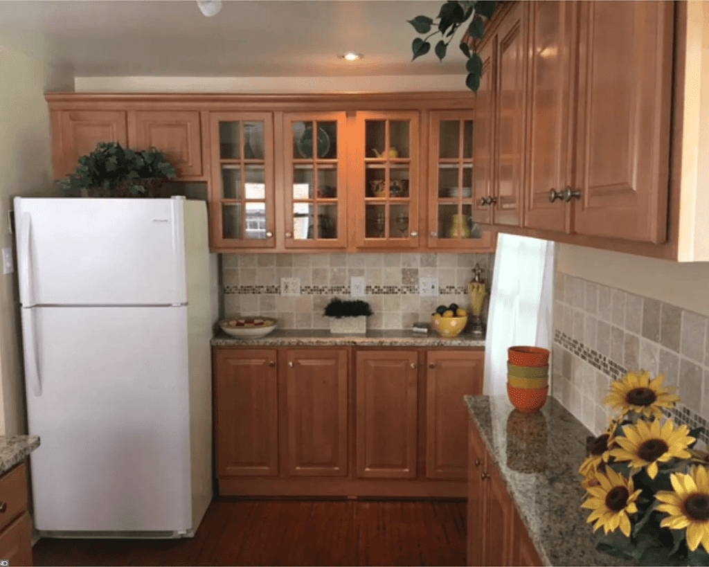 A kitchen with light wood cabinets, granite countertops, a white refrigerator, and a decorative backsplash with mosaic tiles. Sunflowers and plants add color to the space.